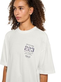 Kobieta o kręconych włosach, ubrana w białą, luźną koszulkę z napisem "OFF DUTY GEAR 223" i małymi nadrukowanymi detalami na piersi.