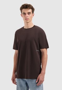 Young man with short brown hair wearing a plain dark brown t-shirt and light blue jeans, standing against a light gray background.