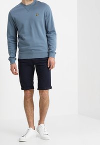 Felpa azzurra chiara con scollatura rotonda e un piccolo logo giallo, abbinata a pantaloni corti blu navy e sneaker bianche. Combinazione di abbigliamento casual.