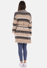 Cardigan oversize a righe beige e nere con orlo a frange e colletto testurizzato, cintrata in vita, indossato sopra jeans blu e scarpe bianche.
