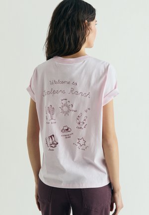 Mujer de cabello largo y oscuro lleva una camiseta rosa claro con las mangas remangadas que tiene el texto "Bienvenido a Calpens Ranch" e ilustraciones de temática occidental.