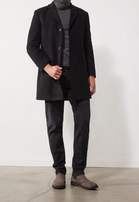 Mężczyzna w czarnym płaszczu, szarym swetrze z golfem, ciemnych spodniach i brązowych zamszowych butach stoi w pomieszczeniu na betonowej podłodze.