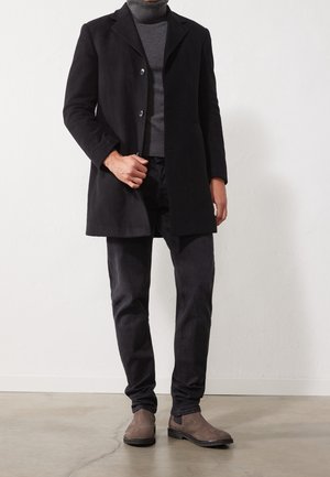 Hombre con abrigo negro, suéter de cuello alto gris, pantalones oscuros y botas de ante marrones, de pie en el interior sobre un suelo de hormigón.