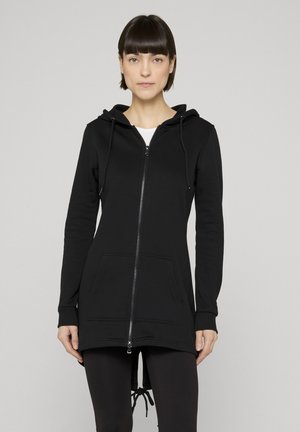 Frau mit kurzen dunklen Haaren, die einen langen schwarzen Kapuzenpullover mit Reißverschluss und Fronttaschen sowie schwarze Leggings trägt, vor einem schlichten hellen Hintergrund.