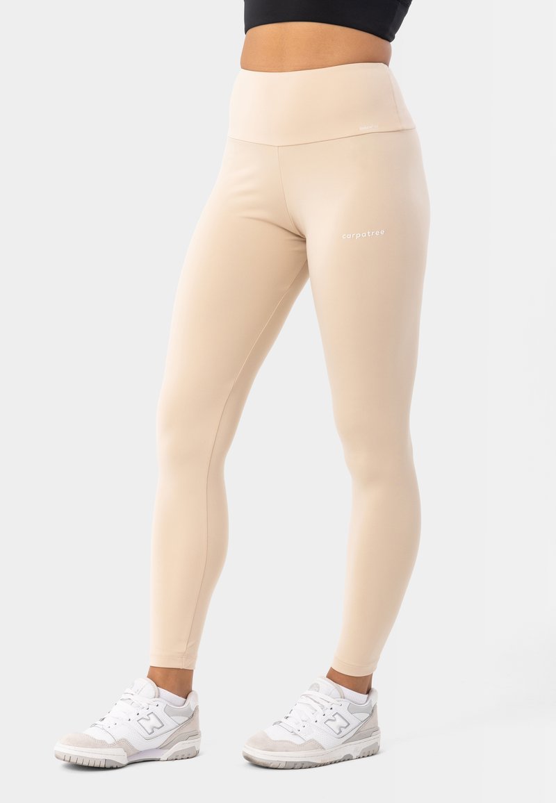 Beige visoko pasne legginge, izdelane iz raztegljivega materiala z gladko teksturo, z majhnim belim logotipom na stegnih in dolžino do gležnjev.