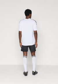 Λευκή αθλητική φανέλα με μαύρες ρίγες στους ώμους, συνδυασμένη με σκούρες σορτς και λευκές κάλτσες μέχρι το γόνατο με μαύρες ρίγες, καθώς και μαύρα παπούτσια.