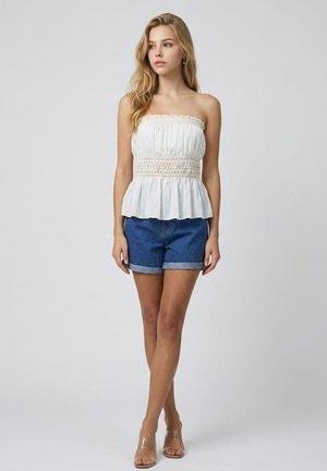 Joven mujer con cabello largo y rubio, que lleva un top blanco sin tirantes con detalles de encaje y shorts vaqueros azules, de pie sobre un fondo liso.