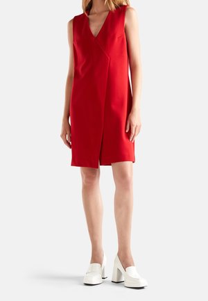 Vestido sin mangas rojo hasta la rodilla con escote en V y solapa frontal, combinado con zapatos blancos de tacón grueso en un modelo de piel clara.