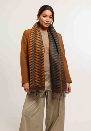 Sciarpa marrone e blu con motivo e estremità frangiate, realizzata in un materiale morbido. Indossata a strati sopra un top beige, abbinata a un cappotto marrone.