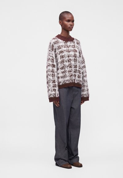 Femme debout portant un pull en maille marron et blanc à motifs, un pantalon ample gris foncé et des chaussures marron sur un fond blanc uni.