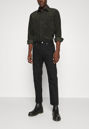 Vaqueros negros de corte slim y diseño de cinco bolsillos, combinados con una camisa de pana verde oscura con botones en la parte delantera y mangas arremangadas.