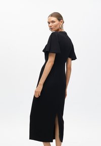 Svart midiklänning med korta utställda ärmar, hög krage och sprund på sidan. Slät tyg med en figursydd silhuett, med dekorativ detalj på baksidan.