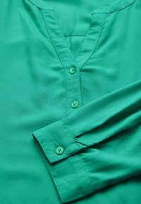 Blusa verde con scollatura a V, chiusura con bottoni e maniche lunghe, realizzata in tessuto morbido, con bottoni verdi coordinati.