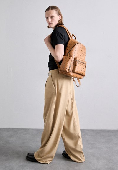 Zaino in pelle tan con motivo, tasca frontale con zip e spallacci regolabili. T-shirt nera e pantaloni beige a gamba larga indossati dal modello.