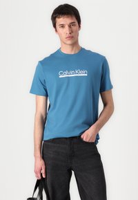 Modrá bavlnená tričko s krátkymi rukávmi a okrúhlym výstrihom, na ktorom je biela loga "Calvin Klein". Skombinované s čiernymi džínami a taškou.