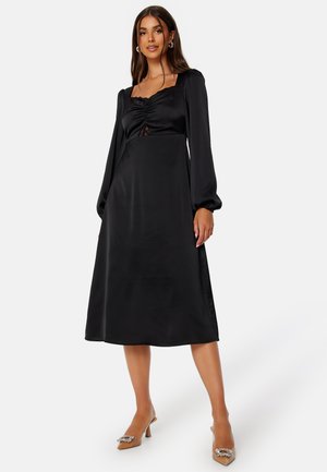 Robe midi en satin noir avec de longues manches bouffantes, un col carré, un corsage froncé et des détails en dentelle. Associée à des chaussures à talons beiges.