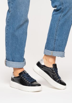 Person wearing blue cuffed jeans and black sneakers with white soles and silver heel accents, standing on a white surface.