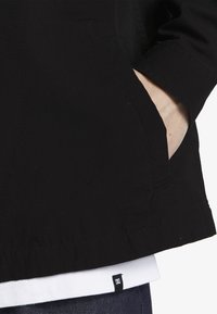 Chaqueta negra con un tejido texturizado, que cuenta con un bolsillo exterior y un detalle de dobladillo en capas en blanco. Pequeño logo de DC visible en el dobladillo.