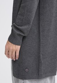 Pilkas megztas drabužis laisvo silueto; turi raukšlėtą rankogalių juostą ir mažą apvalų logotipo pleistrą netoli krašto. Audinys atrodo minkštas ir tekstūruotas.