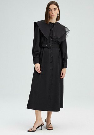 Zwarte jurk met lange mouwen, een grote kraag, gerimpelde accenten, een knoopsluiting aan de voorkant, een taillering met een ceintuur, en zwarte hoge hakken.