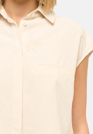 Femme portant une chemise beige sans manches avec col et poche poitrine, montrant le haut du torse et le bras.