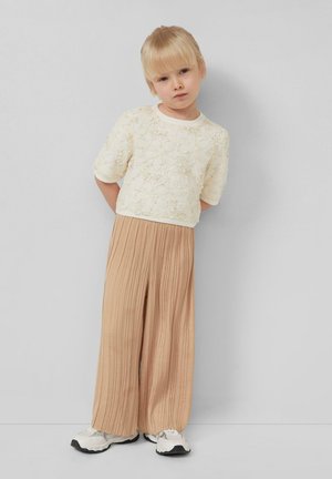 Blondes Kind steht vor einer grauen Wand und trägt ein cremefarbenes Oberteil mit floralem Muster, weite beige plissierte Hosen und weiße Turnschuhe.