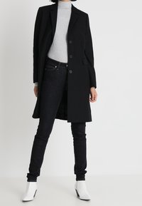 Schwarzer langer Mantel mit einem einreihigen Design und drei Knöpfen, getragen über einem hellgrauen Rollkragenpullover, kombiniert mit dunklen Jeans und weißen Stiefeletten.