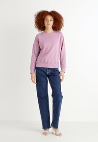 Lilla genser med rund hals og ribbede mansjetter, kombinert med høyveide blå jeans og klare blokkhælede sandaler.