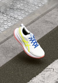 Baskets de course blanches avec des lacets bleus, des accents jaunes et une semelle rose. Présentent un dessus texturé et un design à bout rond.