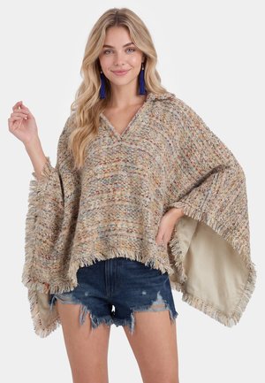Donna bionda che indossa un poncho lavorato a maglia multicolore con frange e orecchini con nappine blu, abbinati a shorts di jeans scuri strappati.
