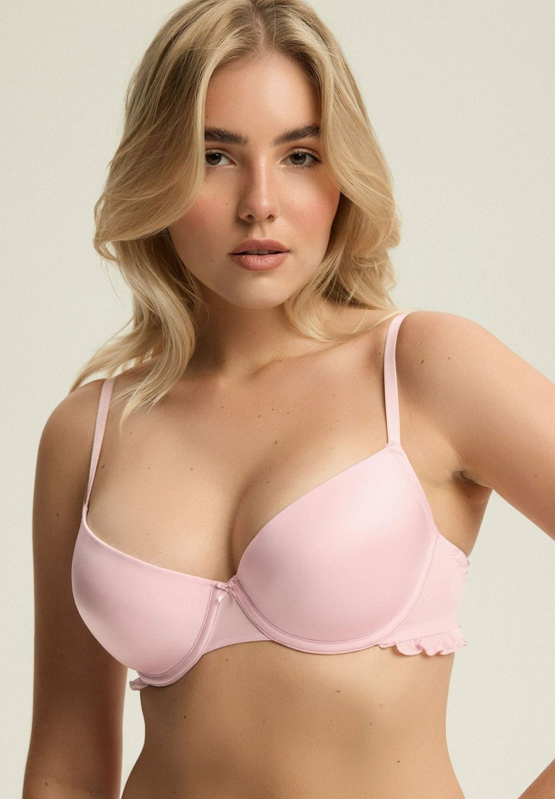 Jasnoróżowy biustonosz z formowanymi miseczkami, cienkimi regulowanymi ramiączkami oraz falbaną na obwodzie, wykonany z gładkiego materiału.