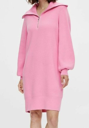 Frau trägt ein rosa gestricktes Langarm-Pulloverkleid mit Reißverschlusskragen und Puffärmeln, steht vor einem schlichten Hintergrund.