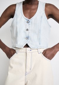 Lys blå denimvest med dyp V-hals og tre metallknapper. Kombinert med kremfargede bukser som har blå sømmer og sidelommer.
