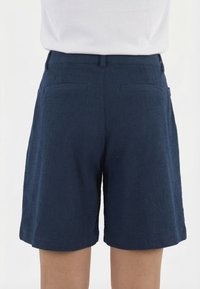 Pantalones cortos azul marino texturizados con trabillas para cinturón y dos bolsillos traseros de ojal, llevados con una camiseta blanca.