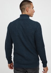 Tmavě modrý pletený svetr s vysokým límcem, strukturovaná látka, žebrované manžety a lem, a raglánové rukávy. Zobrazeno ze zadní strany.