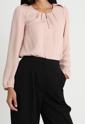 Vrouw draagt een lichtroze blouse met lange mouwen en een frontale rits en zwarte wijde broek met hoge taille met handen in de zakken.