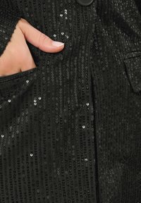 Una mano con smalto rosa chiaro appoggiata nella tasca di un indumento coperto di paillettes nere, con un bottone nero visibile.