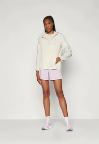 Woman in light beige zip-up jacket, lavender shorts, white socks, and pink sneakers standing against a plain white background.