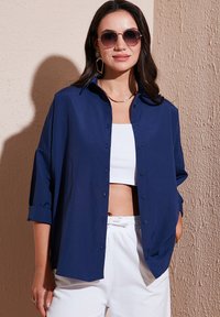 Camicia blu navy con bottoni, realizzata in tessuto morbido, con maniche arrotolate e vestibilità rilassata, abbinata a un crop top bianco e pantaloni chiari.
