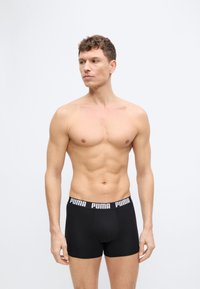 Homme torse nu aux cheveux courts et bouclés portant un boxer Puma noir, debout devant un fond gris clair uni, regardant vers la gauche.