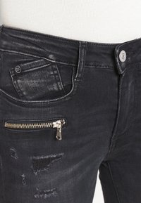 Los jeans ajustados de mezclilla negra presentan una sutil textura desgastada, un bolsillo con cremallera en el muslo y detalles desgastados cerca de la rodilla.