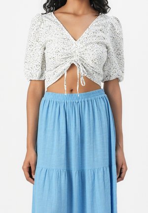 Femme portant un crop top froncé blanc à fleurs avec des manches bouffantes, assorti à une jupe à volants bleu clair taille haute.