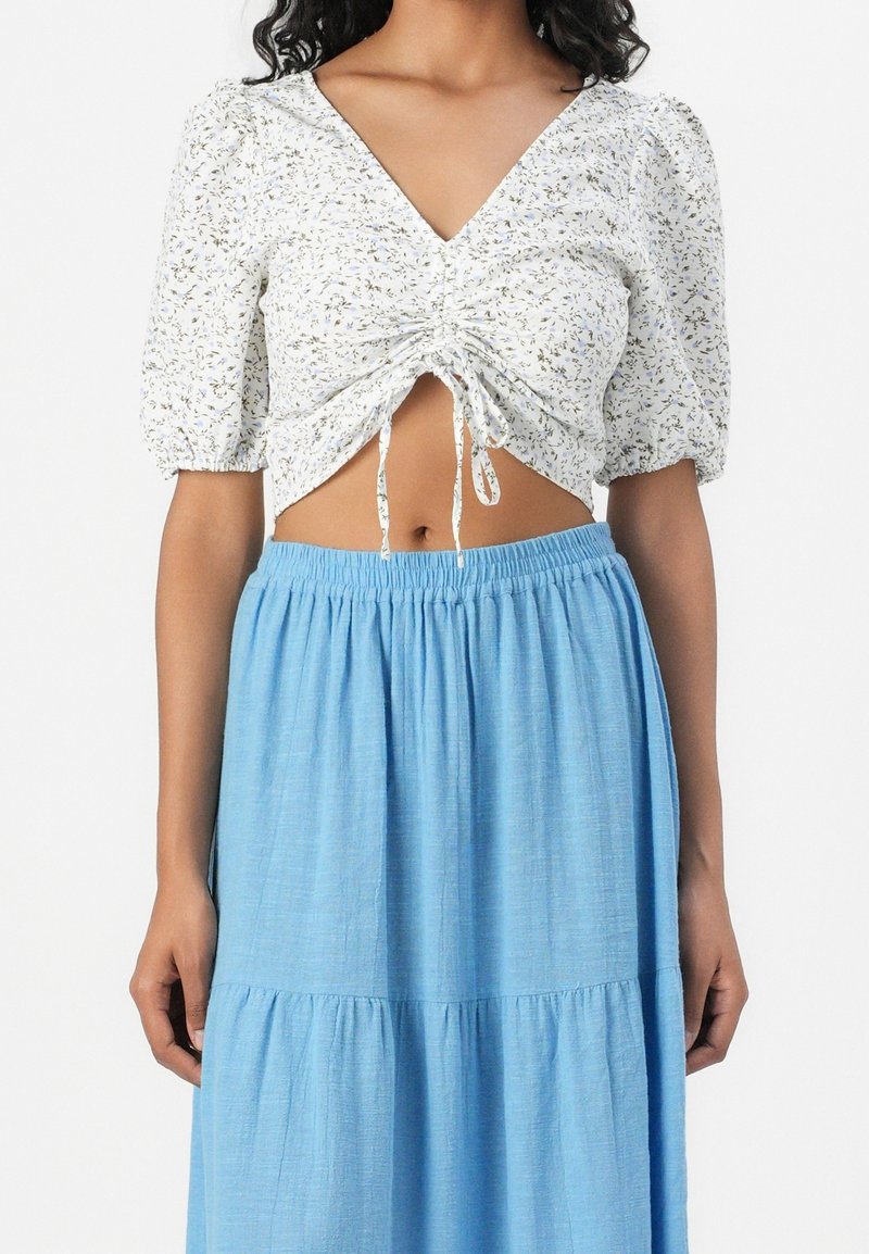 Femme portant un crop top froncé blanc à fleurs avec des manches bouffantes, assorti à une jupe à volants bleu clair taille haute.