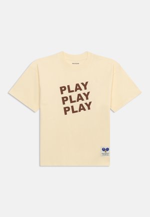 Kortærmet T-shirt i cremefarve med "PLAY PLAY PLAY" i store brune bogstaver på skrå foran og en lille blå lap nær kanten.