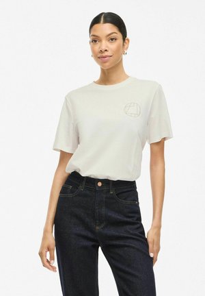 Lysebeige t-shirt med rund halsudskæring og korte ærmer, med et minimalistisk brunt geometrisk logo på brystet, parret med mørke jeans.