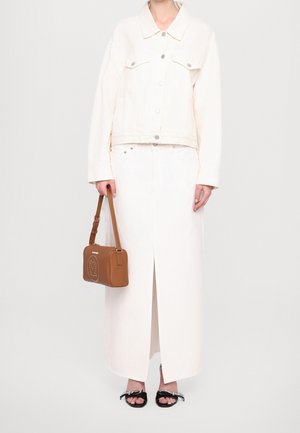 Frau trägt eine cremeweiße Jeansjacke und einen passenden langen Rock mit Schlitz vorne, hält eine braune Handtasche und trägt schwarze offene Sandalen mit Schleife.