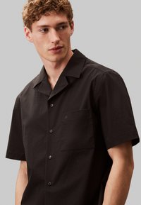 Camisa de botones de manga corta negra con un bolsillo, hecha de un material texturizado. Cuenta con un collar con muesca y una sutil marca en el bolsillo.