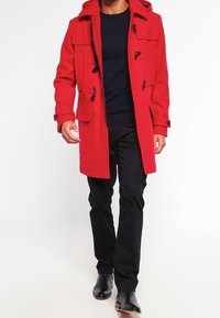 Manteau en laine rouge avec capuche, doté de fermetures à bascule, de deux poches avant et d'une fermeture éclair à l'avant. Porté par-dessus un pull marine et un pantalon noir.