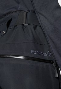 Svarta NORRØNA-byxor med slät textur, som har en ficka med dragkedja, logobrodyr och en justerbar midja. Modern design för utomhusbruk.