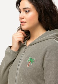 Grå lynlås hoodie med en struktureret vævning, der har en grøn palmetre-patch på brystet og en foret hætte.